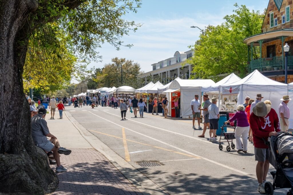 Crowds stroll past white vendor tents lining the downtown street at the Fairhope Arts and Crafts Festival 2026 with a historic Victorian building in the background