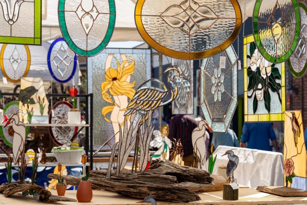 Handcrafted stained glass panels with herons and floral designs displayed alongside driftwood sculpture at a vendor booth during the Fairhope Arts and Crafts Festival 2026